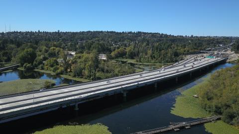 The west approach bridge north, a wide concrete highway bridge over water, stretches left to right. In the background is a neighborhood with lots of tree cover.