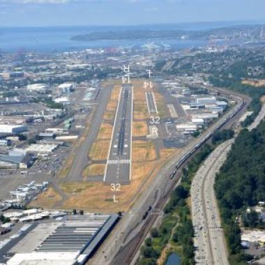 The Boeing Field Runway is shown, featuring two landing strips tucked between an industrial area and a freeway. A beautiful blue sky sits above the surrounding water in the background of the photo. 