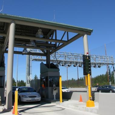 A sedan stops at a toll booth to pay a toll, visible in the background is the main lanes of the roadway with tolling equipment mounted on the gantries over the roadway. 