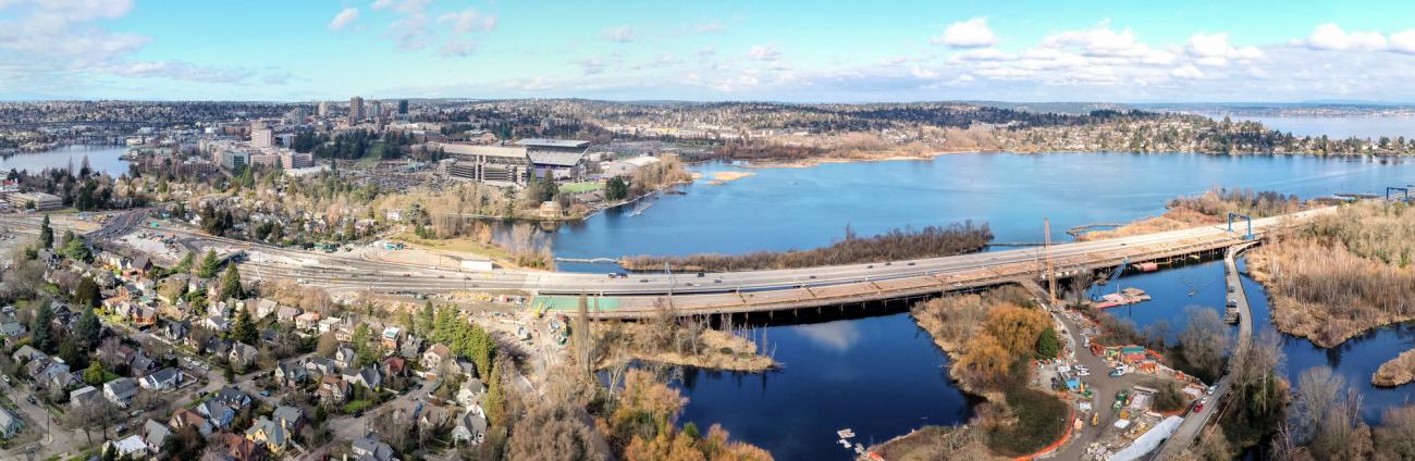 Panorama view of SR 520 across Union Bay. In the foreground is a work zone where crews are construction a new, parallel bridge. The highway is surrounded by a residential neighborhood.