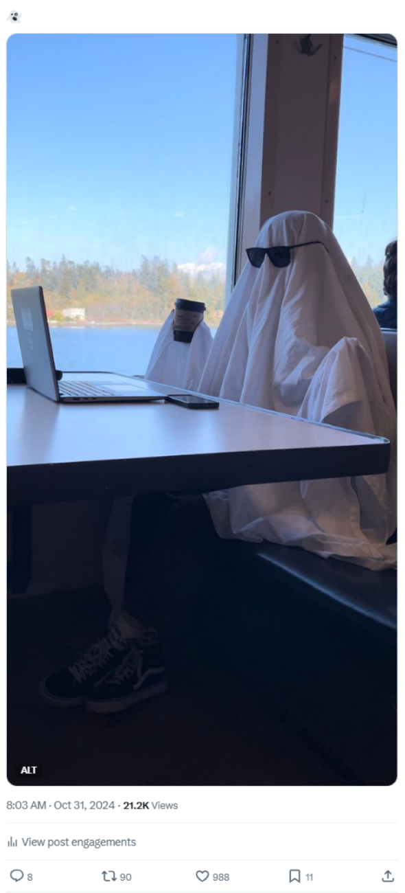 Screenshot of an Oct. 31 X post of a ghost emoji and a photo of a person dressed in a white sheet resembling a ghost, sitting in the passenger cabin of a ferry next to a window. The person is wearing black sunglasses and holding a coffee cup in one hand. On the table in front of them is a laptop and a smartphone.