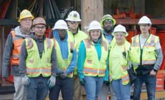 A group of seven people of various ages, races and genders stand in construction vests and hard hats and smile at the camera.