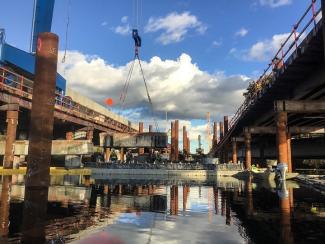 In a construction zone over water, a section of concrete is lifted by a crane. On either side are two tall wooden bridges.