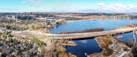 Panorama view of SR 520 across Union Bay. In the foreground is a work zone where crews are construction a new, parallel bridge. The highway is surrounded by a residential neighborhood.