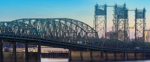 Steel truss bridge over a river with a cityscape background during a sunset or sunrise, and the IBR in the top right corner.