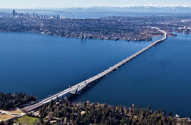 Photo of the new SR 520 floating bridge, looking west across a blue Lake Washington, with the Medina shoreline in the foreground, and the downtown Seattle skyline in the distance, at upper-left.