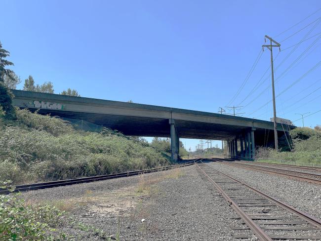 A highway bridge stretches over railroad tracks. A blue sky is visible above and light green vegetation surrounds the short hillsides alongside the bridge.
