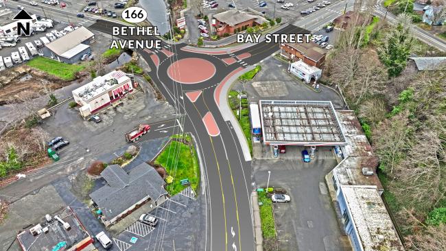 Bird's eye view of the intersection of Bay Street and Bethel Avenue in downtown Port Orchard  with design visual overlay of new single-lane roundabout. New roundabout features raised portions of the roadway to steer vehicles into the roundabout and provide access for bicyclists and pedestrians. Local businesses adjoin intersection, parked vehicles and overhead utility lines. 