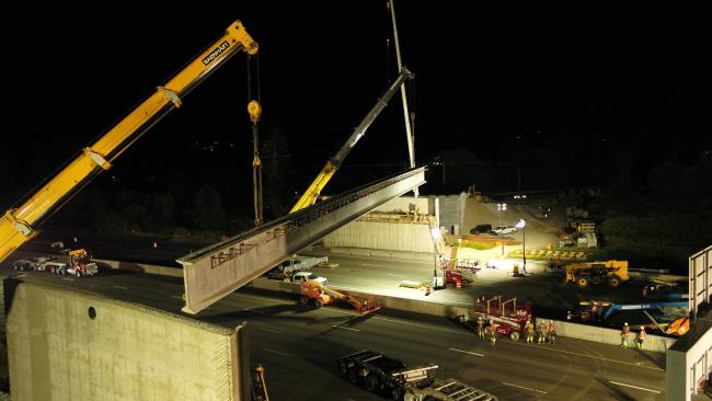 A photo taken at night, showing two large construction cranes lifting the first Wapato Way East bridge girder into place