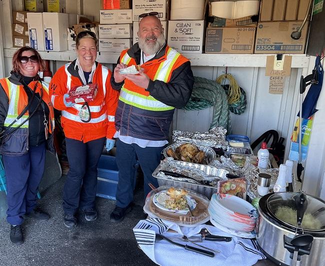 Three people in safety vests enjoy a meal alongside a table full of various Thanksgiving dishes