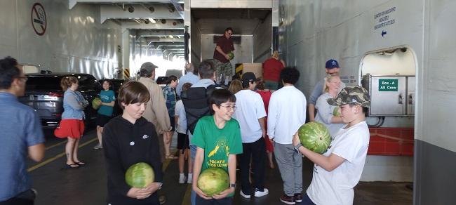 Several people holding watermelons as a person hands them out from a truck on the car deck of a ferry
