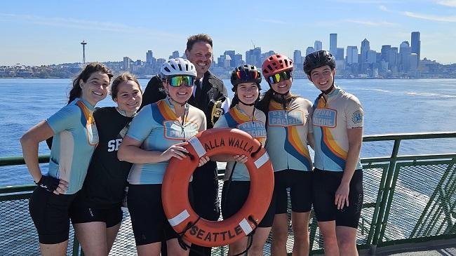 Six bicyclists posing for a photo with a ferry captain and life ring on the outdoor deck of vessel with Seattle skyline in the background