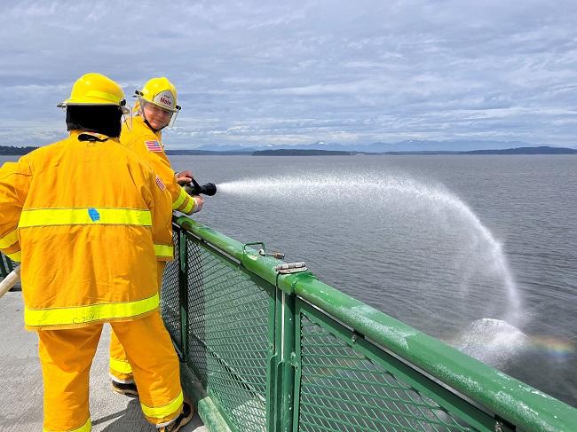Two people in firefighting gear spraying water off the side of a ferry's outdoor deck
