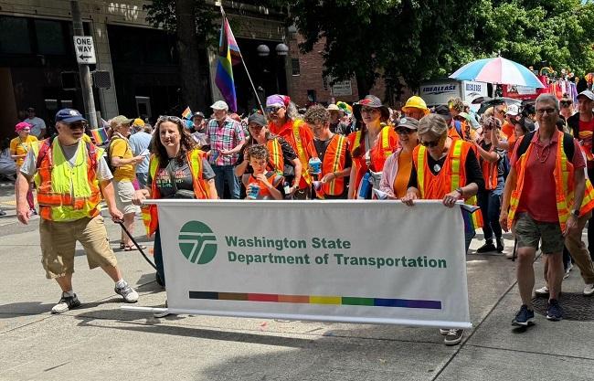 Several people in orange safety vests walking down a city street holding a WSDOT banner