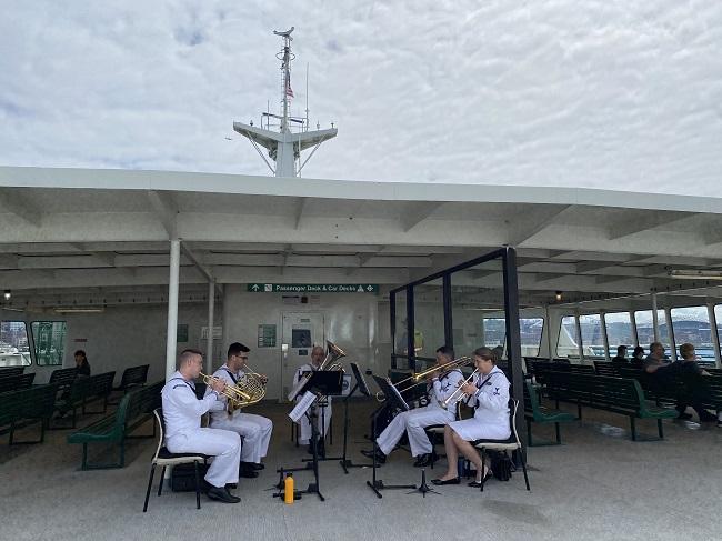 Five people seated in Navy uniforms playing a brass intrument on the sun deck of a ferry