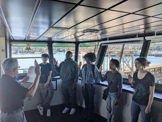 Several people in the wheelhouse of a ferry raising their right hand up