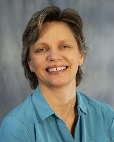 A smiling headshot of Julie Meredith, the WSDOT secretary of transportation. She is wearing a light blue shirt and has short dark hair, she is framed by a light grey background.