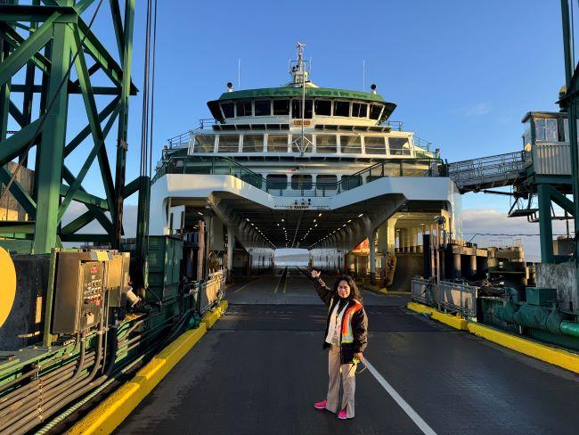 A woman stands in front of a large docked ferry, pointing towards it on a sunny day