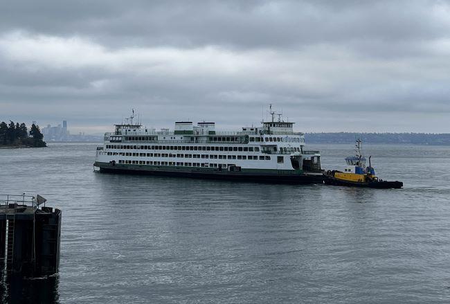 A ferry and a tugboat on a body of water with a distant city skyline under a cloudy sky