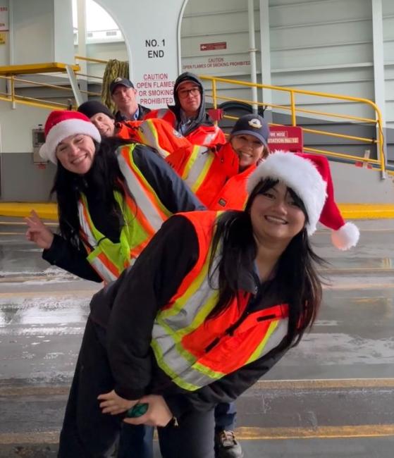 Five people in safety vests, two with Santa hats, standing and smiling on the car deck of a ferry