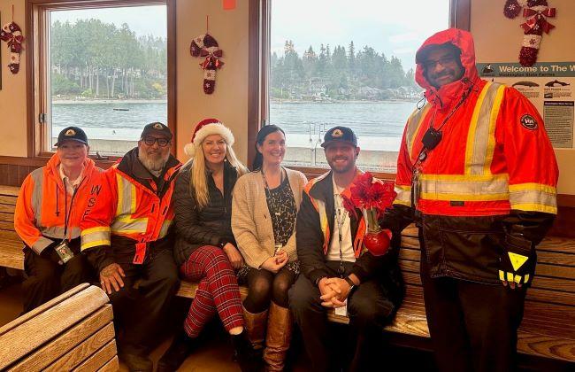 A group of six people, some in orange safety jackets, sit on a bench inside a building with a water view and holiday decorations