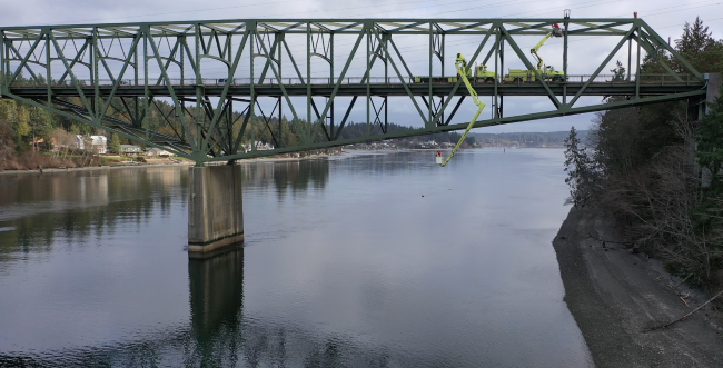 A green metal truss bridge over calm water with maintenance vehicles on top and forested surroundings