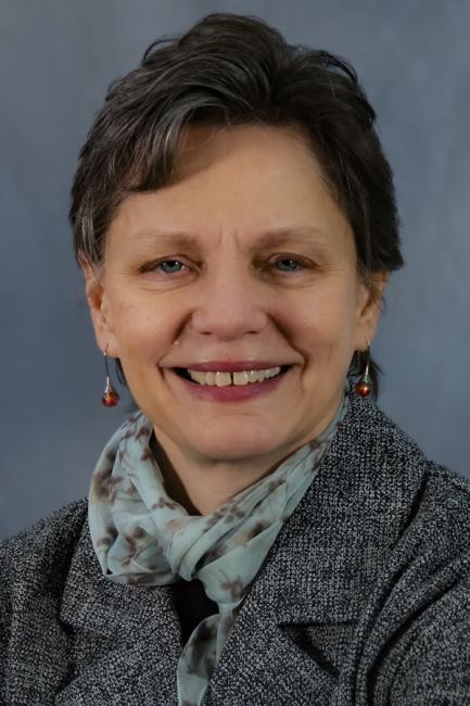 A smiling headshot of Washington State Secretary of Transportation Julie Meredith. She is wearing a gray jacket and has short dark hair. She is framed by a blue background.