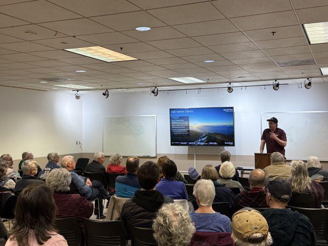 A speaker addresses an audience seated in a meeting room with a screen displaying "Oak Harbor Library"