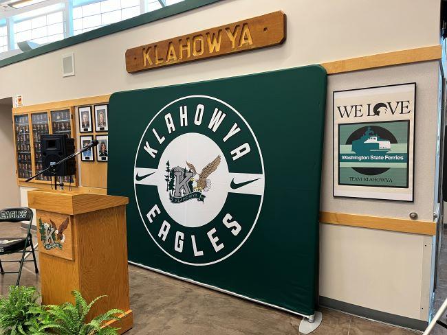 Indoor setting with a green backdrop featuring "KLAHOWYA EAGLES" emblem, a boat's nameplate "KLAHOWYA," a wooden podium and a poster about Washington State Ferries