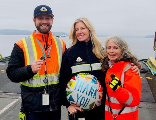 Three people in safety attire posing on a dock with a "THANK YOU" balloon and two of them each holding up a coin